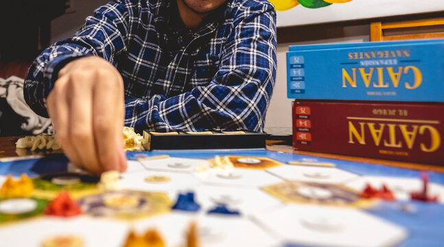 Closeup of beautiful white hand holding a 'Catan' board game piece (from 'Seafarers' expansion) and young man with shirt and faceless with interior light