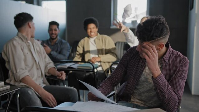Nerd student sits in classroom at desk alone reads summary suffers from cruel aggressive attitude from classmates young guy loser feels victim of bullying experiencing frustrated resentment depression