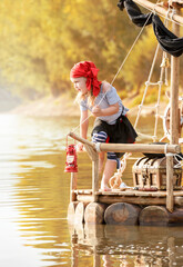 Child in a pirate costume plays on a wooden raft at sunset. Girl pretends to be the captain of a...