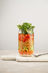 Trendy Fresh detox summer salad layered in a glass mason jar: yoghurt dressing, carrots, cucumber, yellow lentils, red bell pepper, green leaves and herbs, long silver spoon, on white background