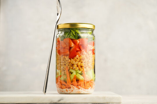 Trendy Fresh detox summer salad layered in a glass mason jar: yoghurt dressing, carrots, cucumber, yellow lentils, red bell pepper, green leaves and herbs, long silver spoon, on white background