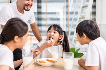 Asian  family enjoying breakfast at living room. little girl daughter sitting on table, drinking milk with smiling father and mother in morning. Happy family at home.