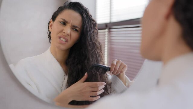 Close-up young woman looking in mirror combing hair in bathroom annoyed sad girl dissatisfied damaged dry tangled hair condition frustrated about split ends vitamin deficiency morning daily routine