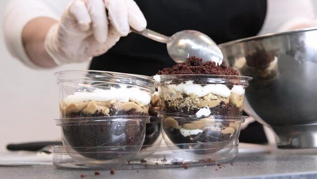 Woman cook in white gloves sprinkles a layered dessert in a transparent form with a biscuit from above. Slow motion video