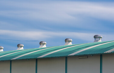 Round stainless steel roof ventilators on corrugated metal rooftop of industrial building against clouds on blue sky background