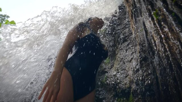 A slender girl in blue swimsuit stands under flow of a tropical waterfall waterfall. a sexy girl stands under a rock, from which a stream of a mountain river flows down on a sunny day. Slow motion