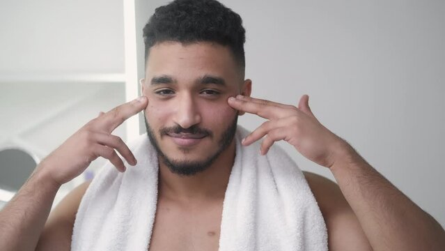 Anti-aging skincare. Facial moisturizing. Morning grooming. Confident satisfied man applying face cream under eyes at light bathroom interior.