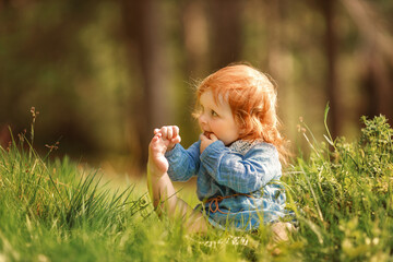 Funny red hair baby is sitting on the grass and holding her own leg like yoga. Cute child putting...
