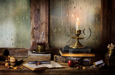 Classic still life with illuminated candle placed with vintage books, hot drink, sugar, ,old boxes, pipe, nuts and nutcracker on a rainy day window background.