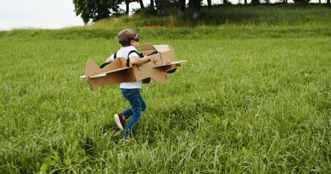 Happy child playing outside on green grass. Kid pilot with a toy paper plane. Kid boy play with toy plane cardboard. Success winner, imagination and dream concept. High quality 4k footage
