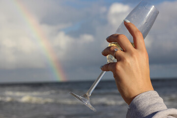 hand with engagement ring holding champagne with beach rainbow background