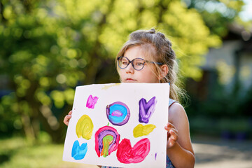 Little preschool girl holding picture painted with colorful water colors. Happy child with glasses, education and school concept. Creative leisure for children.