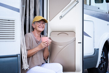 Smiling senior woman in travel vacation leisure outside a camper van enjoying freedom. Attractive elderly caucasian lady in van life sitting at the door of motor home with a coffee cup looking away.