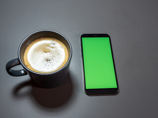 A cup of coffee and a phone with a green screen on a gray table.