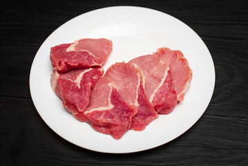 Fototapeta premium Raw pork chop on a white plate on a black wooden background. Fatty meat beaten off for cooking.
