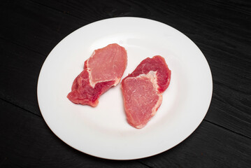 Raw pork chop on a white plate on a black wooden background. Fatty meat beaten off for cooking.
