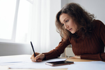 Wide angle shot of pensive serious focused curly woman writing script counts taxes calculates...