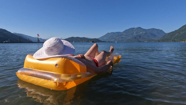Mid adult woman lying on inflatable mattress, wearing a white straw sun hat that obscure her face while floating on the beautiful Harrison Lake surrounded with mountains in Harrison Hot Springs