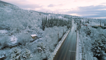 Aerial for snowy forest and a and car moving on the winter road. Shot. Aerial view of the road through a winter forest with a moving vehicle on cloudy sky background.