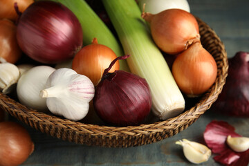 Wicker basket with fresh onion bulbs, leeks and garlic on light blue wooden table, closeup