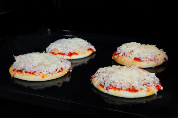Four mini pizzas with ham, cheese and sause baking on tray in electric oven, black background. Italian cuisine, homemade bakery, fast food, cooking concept