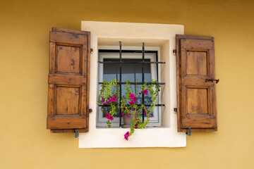 A beautiful typical Italian window with steel bars, brown wooden shutters and a purple petunia plant.