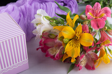 Greeting concept. Bright yellow, pink, white flowers lay near striped present box and purple fabric. Fresh alstroemeria lilly bouquet