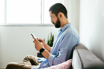 Young brazilian man sitting on sofa using smartphone