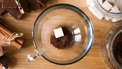 Top view of instant coffee preparation process. Concept. Putting coffee, sugar cube, and hot water into the transparent mug on wooden table background with chocolate and cinnamon.