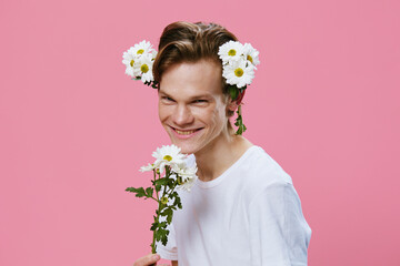 a cute, handsome guy in a white t-shirt smells a bouquet of large chamomile flowers in his hands, and there are also flowers in his hair.