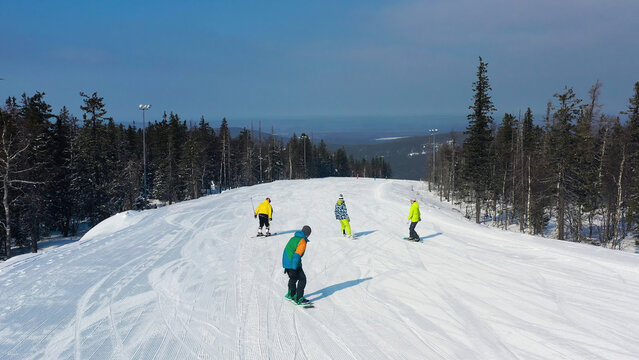 Modern Ski Resort In Pine Forest Aeria, View From Above. Footage. Young Group Of People Snowboarding And Skiing Down The Snowy Slope.