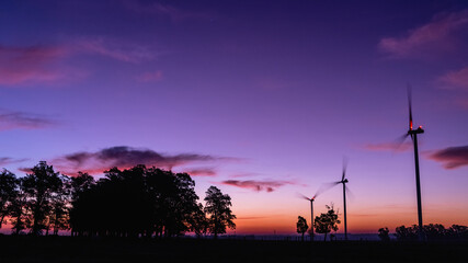 The silhouette of three modern windmills in a colorful sunset, next to a group of trees on the horizon, on the outskirts of Kiyú, San José, Uruguay