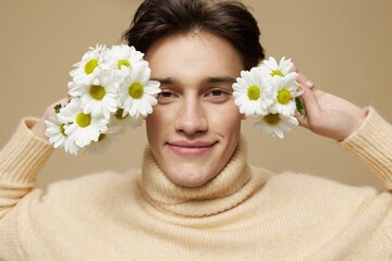 Obraz na płótnie Canvas A handsome man with light white skin, with dark, short, red hair combed back, in a beige turtleneck with a high collar stands on a dark beige background with a bouquet of daisies near his face.Close