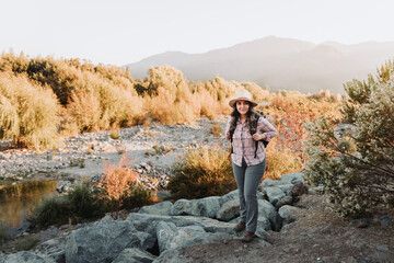 Fototapeta premium Smiling young latin woman with a hat and a backpack on, hiking in a beautiful landscape.
