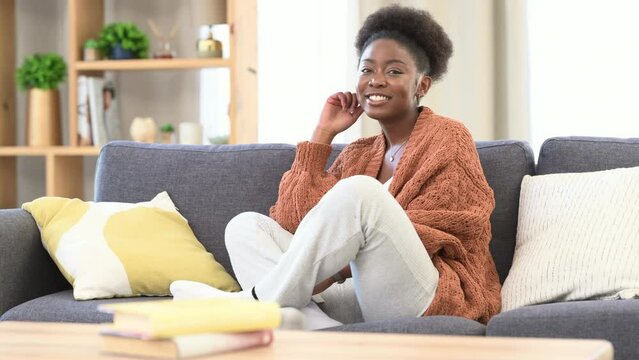 Happy woman looking at camera smiling on sofa. Lady with big smile relaxing in home environment, cosy and warmly sitting on couch feeling safe. Female surrounded by comfort in a light radiant lounge.