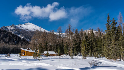 Wooden houses stand on the edge of a coniferous forest. There is a man standing next to a snowmobile in a snowy valley. Picturesque mountains against a background of blue sky and clouds. Altai