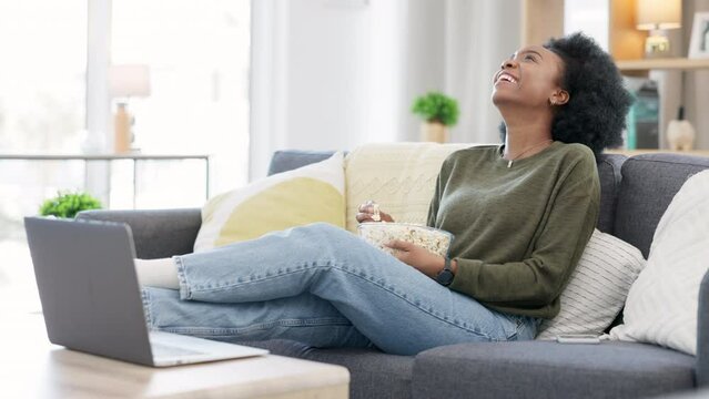 Happy african american woman streaming online movies on a laptop while snacking on popcorn and relaxing on a sofa at home. Black female enjoying a comedy, eating, laughing and having fun on a weekend
