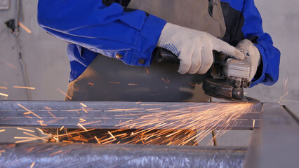 Craftsman sawing metal with disk grinder in workshop. Grinding metal with sparks flying. Electric wheel grinding on steel structure in factory Low speed shutter. Metal grinding with orange flying