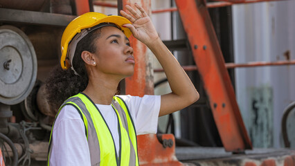 Young African American mechanical female engineer in safety suit and yellow helmet stands next to...
