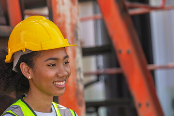 Young African American mechanical female engineer in safety suit and yellow helmet stands next to...