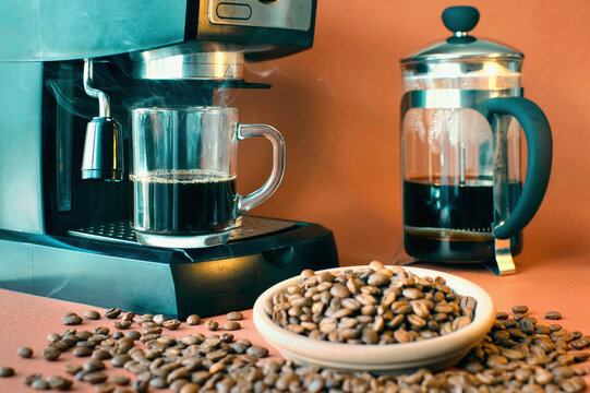 Closeup Shot Of Expresso Maker, Glass Coffee Mug And Spilled Coffee Beans In Front 