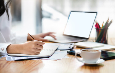 Close up view, young asian businesswoman sitting on desk in office writing her plan on note book, use computer laptop working with paperwork of investment, select focus on hand holding pen