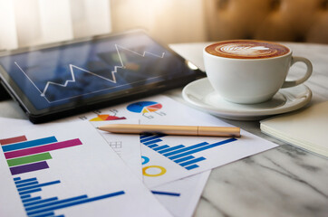 Close up view, latte coffee in white cup with pen, chart of investment and tablet computer on table in cafe, business people take a break during a day in cafe and working part time