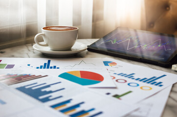 Close up view, latte coffee in white cup with chart of investment and tablet computer on table in cafe, business people take a break during a day in cafe and working part time