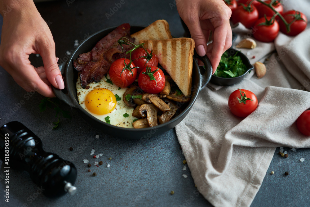 Wall mural Fried eggs, bacon, tomato and toasted bread in a black ceramic pan at domestic kitchen