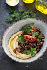 Hummus served with ground beef meat, fresh parsley and tomato wedges in a grey bowl, middle close-up on a brown stone background