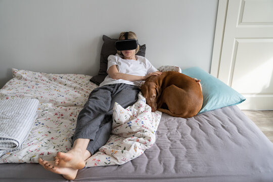 Woman wearing VR helmet. Middle-aged female in virtual reality goggles resting at in relaxing virtual environment at home near her dog, lying at the bed. AR, cyberspace concept