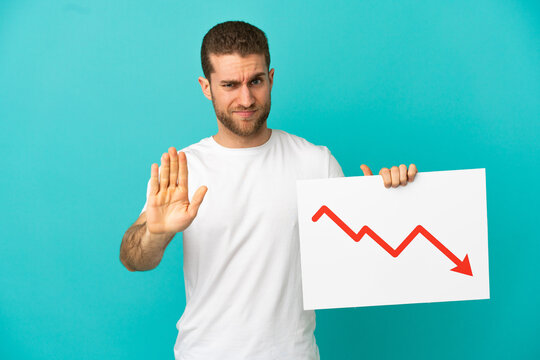 Handsome blonde man over isolated blue background holding a sign with a decreasing statistics arrow symbol and doing stop sign