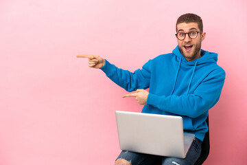 Young man sitting on a chair with laptop surprised and pointing side
