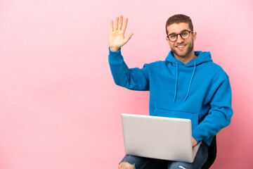 Young man sitting on a chair with laptop saluting with hand with happy expression